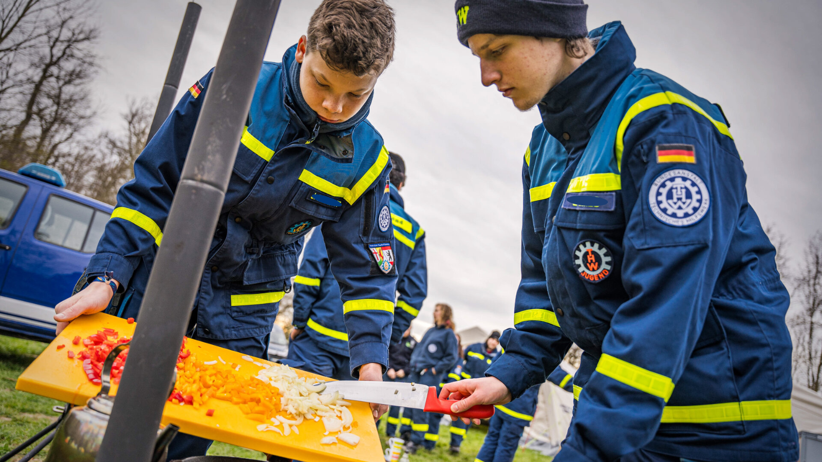 Zwei Jungen in Uniform der THW-Jugend bereiten auf einer Campingwiese Gemüse auf einem Schneidebrett zu.